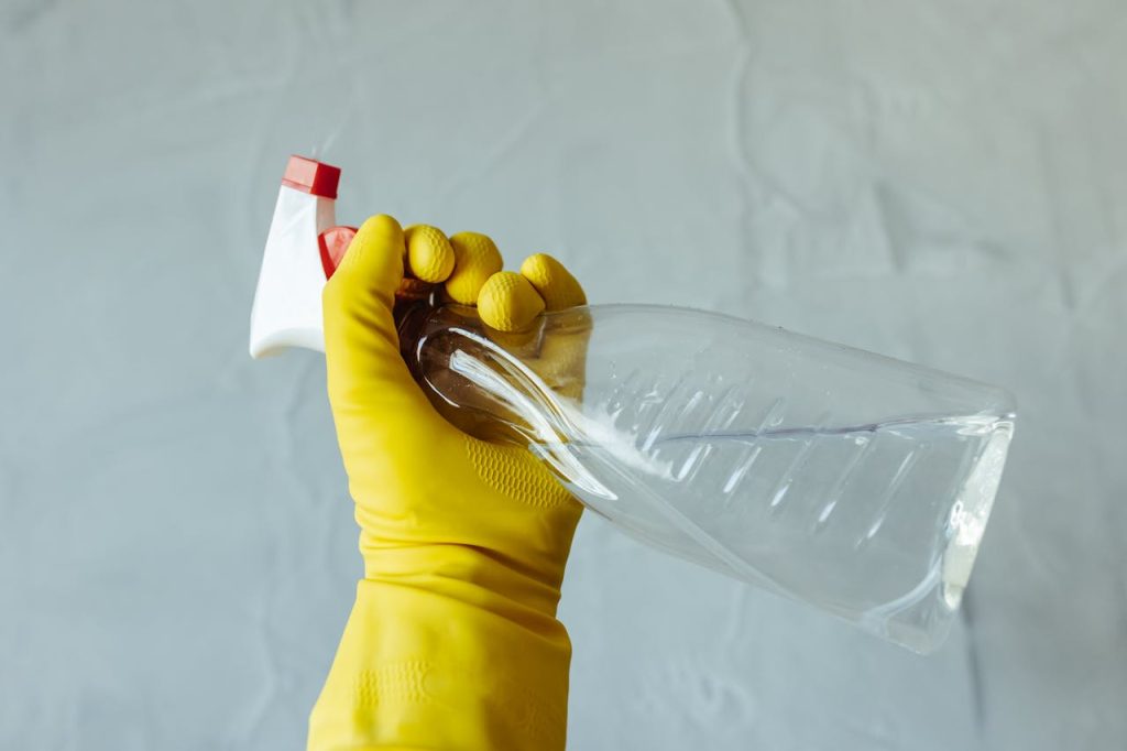 pexels-photo-28576634 Close-up of a hand wearing yellow gloves holding a transparent spray bottle, isolated on a plain background.